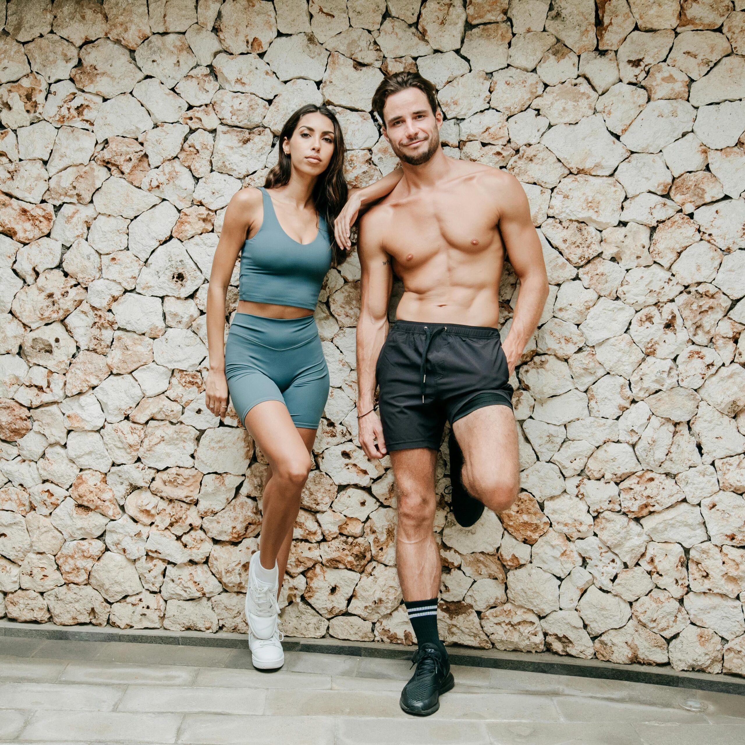 A fit couple in sportswear posing confidently against a textured stone wall outdoors.