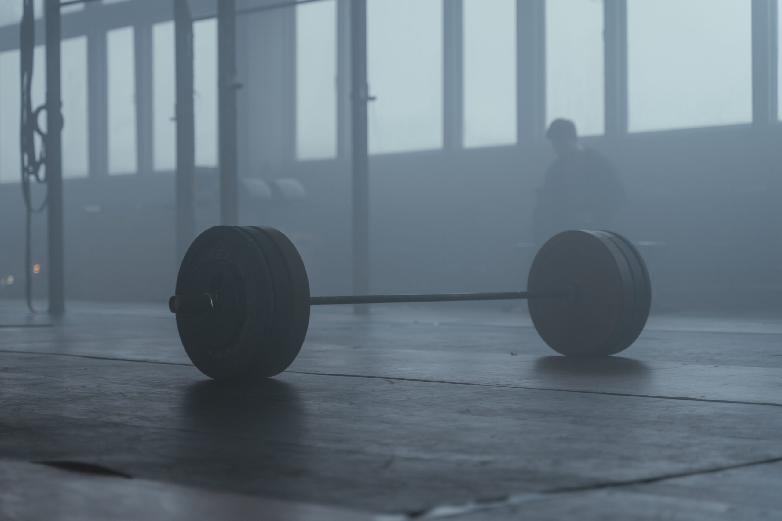 A barbell loaded with large weight plates rests on a wooden gym floor, photographed in a hazy, fog-filled industrial space with tall multi-pane windows and the blurred silhouette of a person standing in the background.